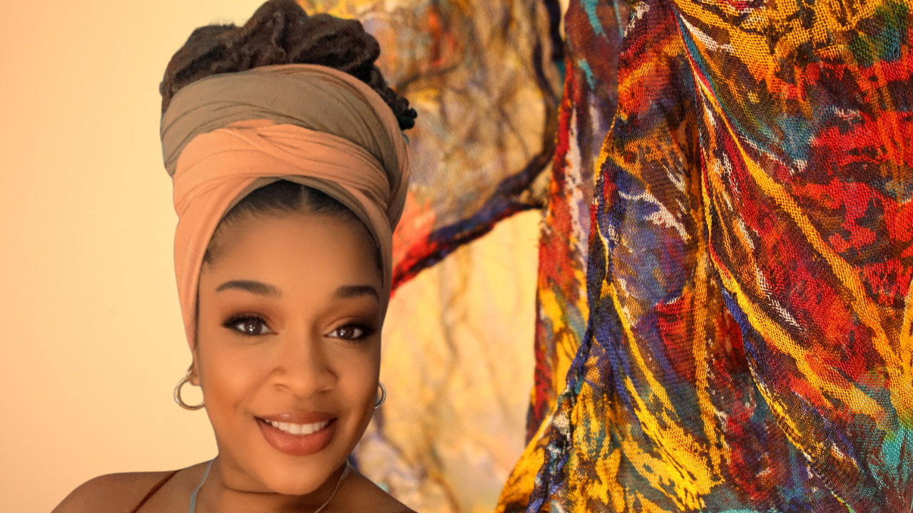 Thiy Parks wears a tan and olive green headwrap atop a bun of brunette locs for National Headwrap day. Behind her is colorful draped fabric.